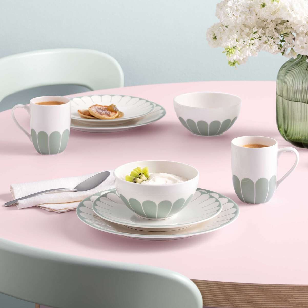 Set di stoviglie in ceramica bianca con decorazioni verdi, secondo piatto, ciotola con yogurt e kiwi, tazza con caffè, su tavolo rosa.