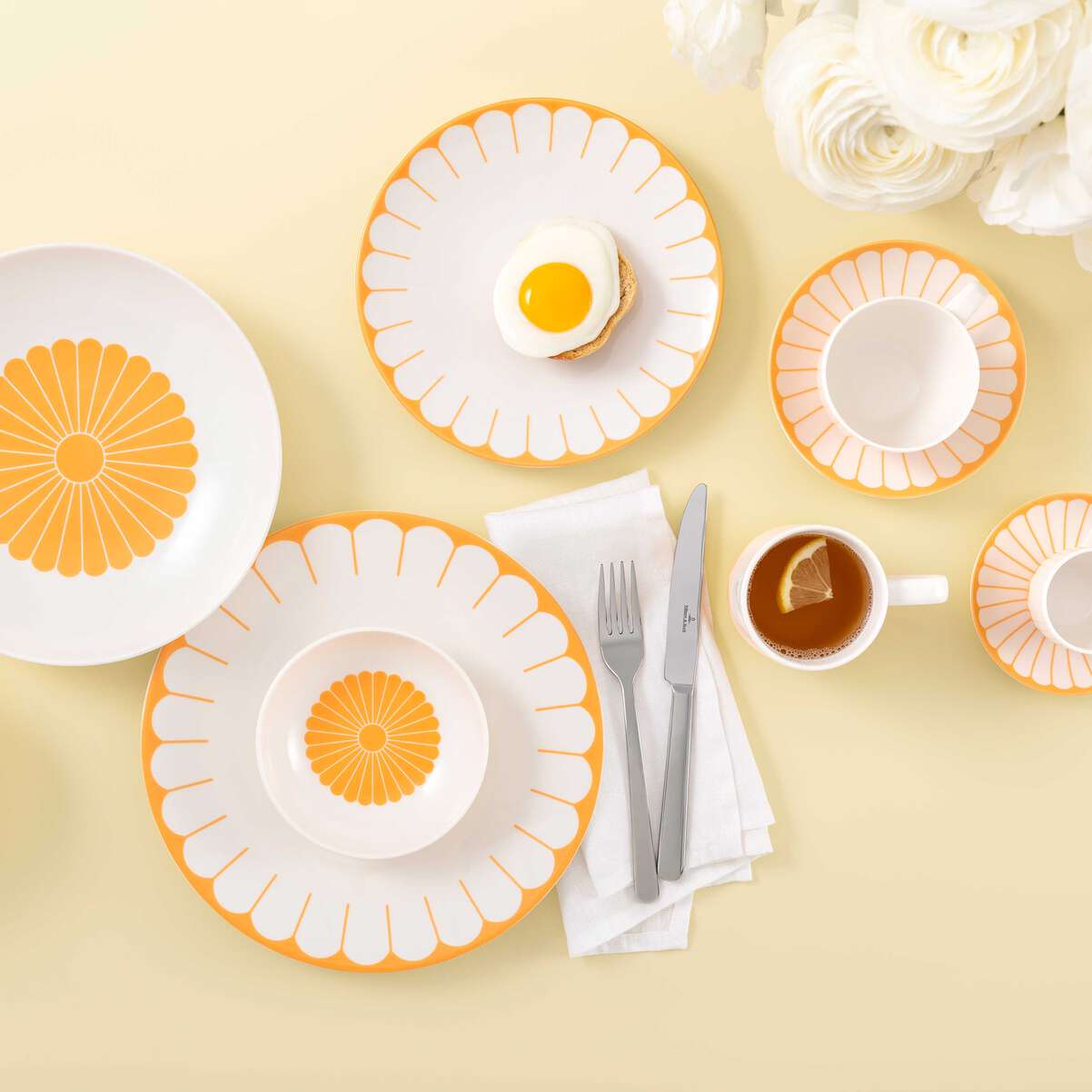 Set da tavola con piatti bianchi e arancioni, posate grigie, una tazza di tè e un piatto con un uovo all'occhio di bue su un sottofondo giallo.