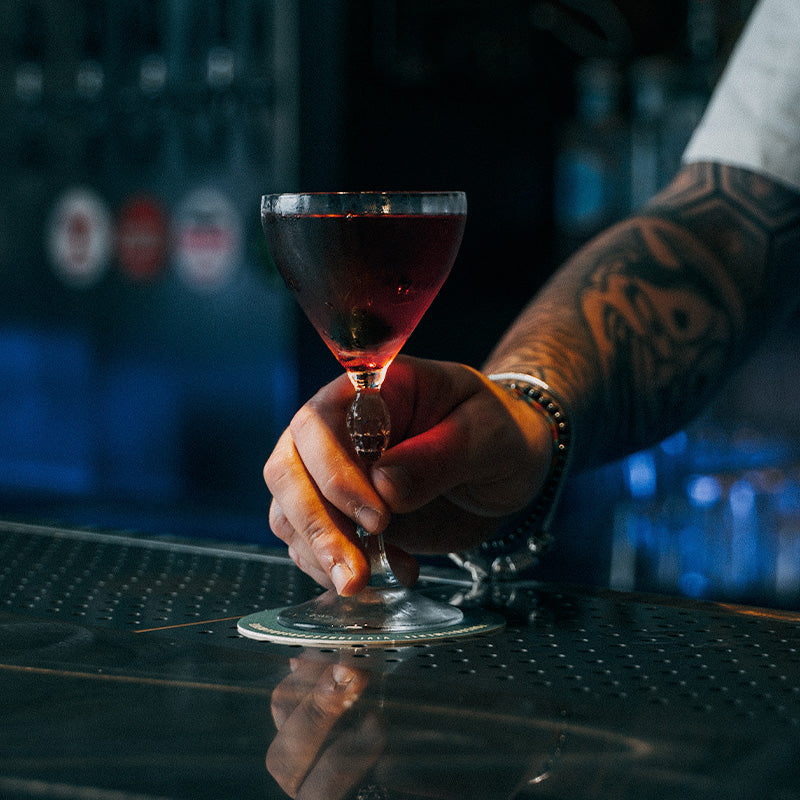 mano con braccialetti e tatuaggi che regge un bicchiere da cocktail con liquido rosso scuro su bancone metallico riflettente