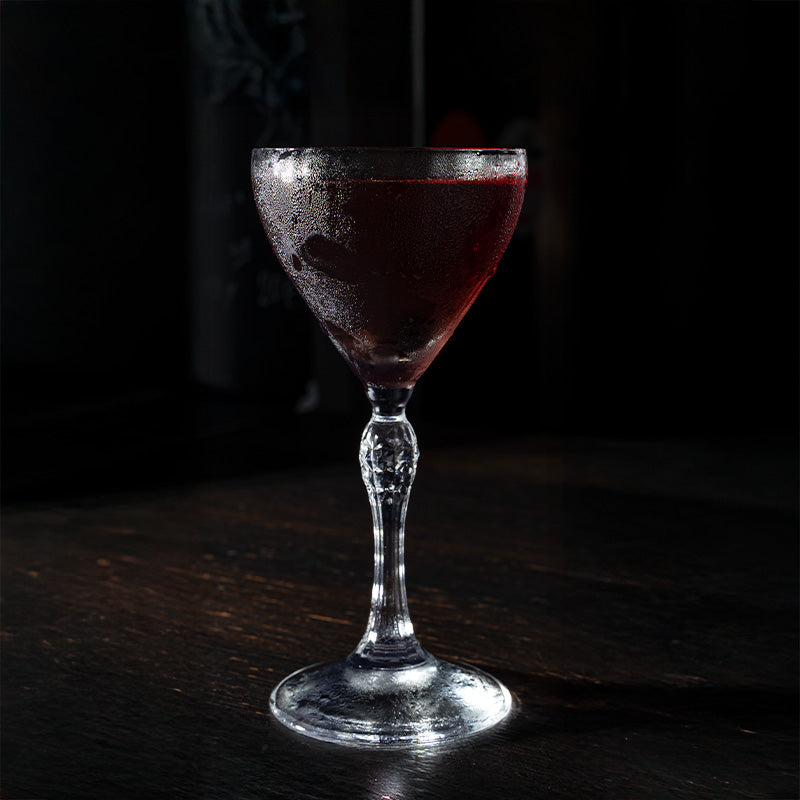 Calice di vetro trasparente con gambo decorato, contenente liquido rosso scuro condensato, su tavolo scuro e sfondo nero.