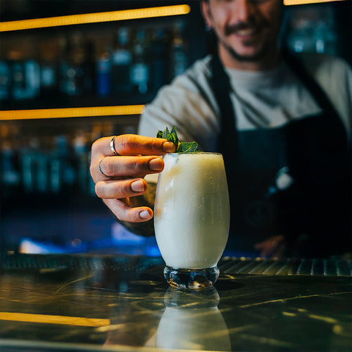 Bicchiere di vetro trasparente con cocktail bianco e foglie di menta, mano con due anelli lo sta posando su bancone riflettente in bar.