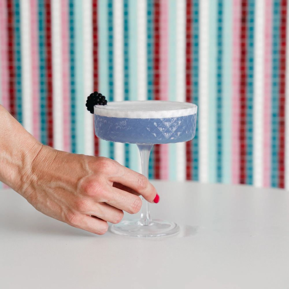 mano con smalto rosa che tiene calice di vetro con cocktail viola con schiuma bianca e mora su sfondo a righe colorate