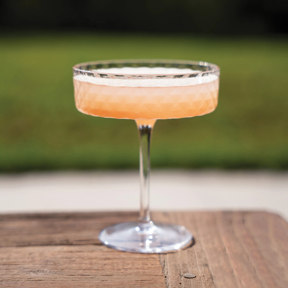 Calice trasparente con superficie sfaccettata, contenente un cocktail rosa tenue, appoggiato su tavolo in legno, sfondo sfocato verde.