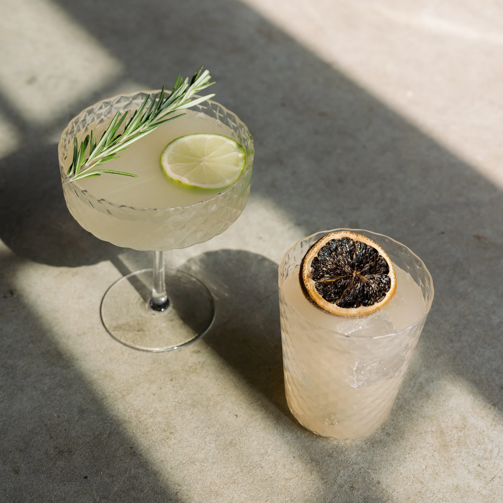 Due cocktail chiari in bicchieri di vetro lavorato, uno con fetta di lime e rametto di rosmarino, l'altro con fetta di agrume essiccato, su