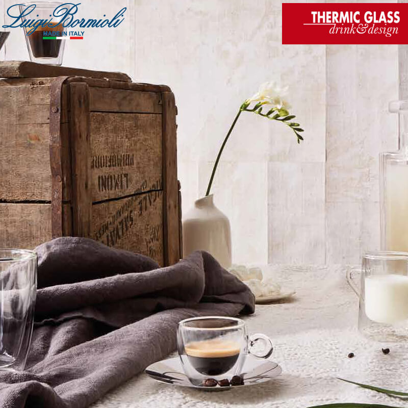 Transparente Doppelwand-Glas-Tasse mit Kaffee auf silberner Untertasse, grauer Tischdecke, weißer Vase und Holzkiste