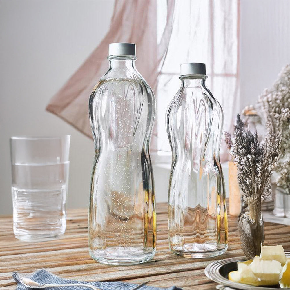 Due bottiglie di vetro trasparente, una con bollicine, accanto a un bicchiere d'acqua e un piatto con burro. Ambiente luminoso e caldo.