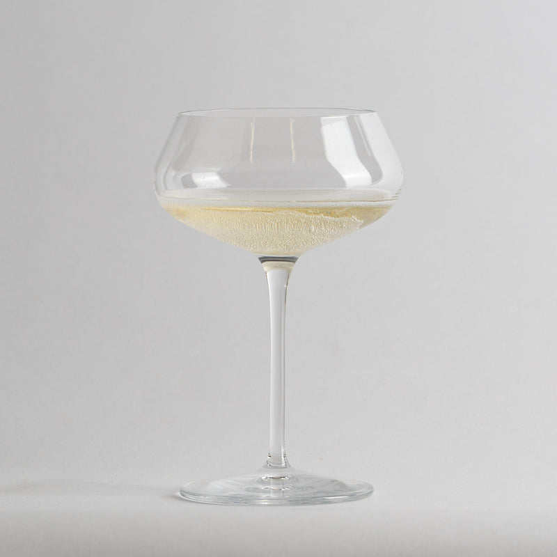 Calice di vetro trasparente a coppa con vino bianco frizzante su sfondo grigio chiaro.
