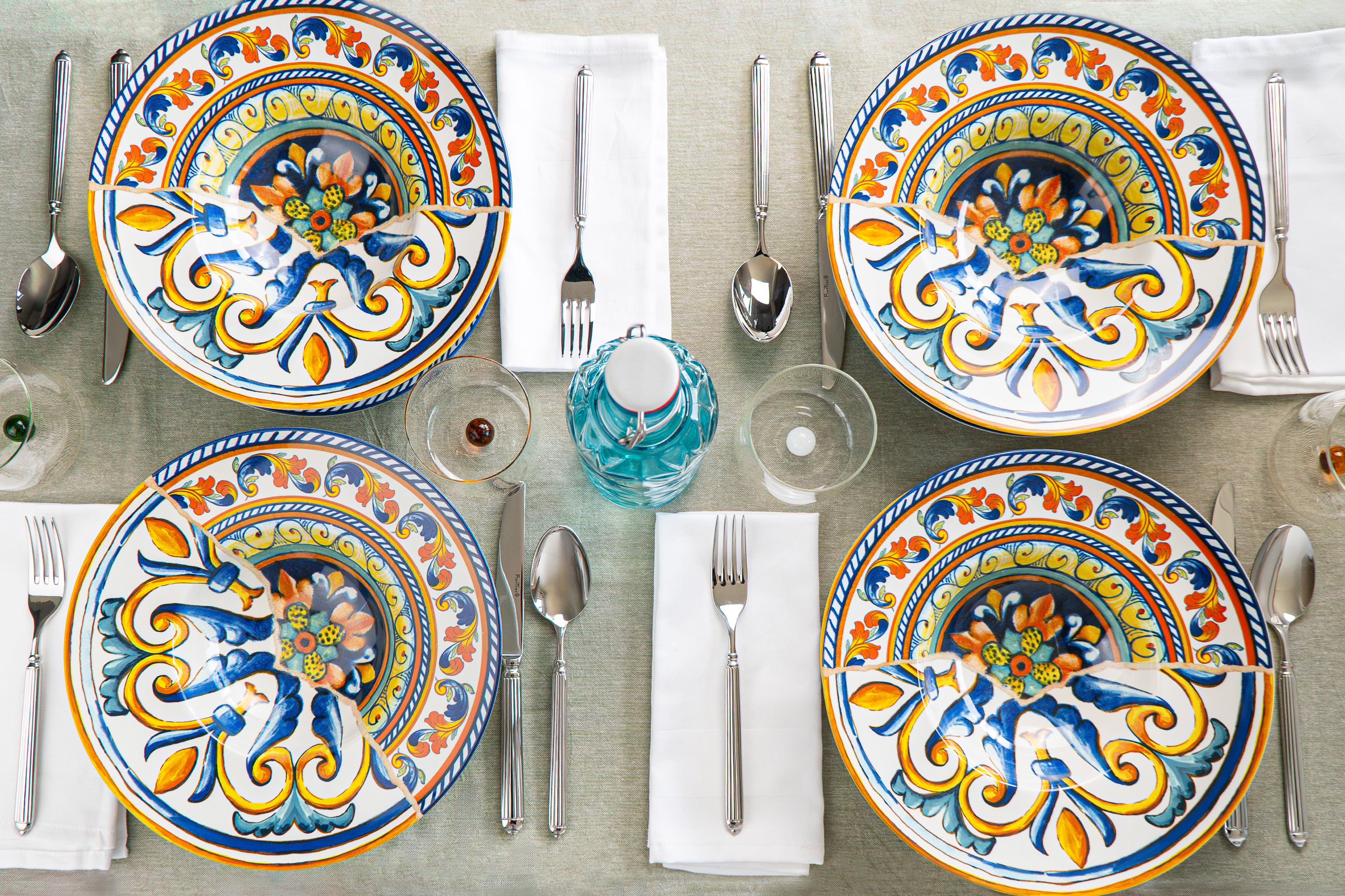 Set di piatti decorati con motivi colorati, posate in acciaio, bicchieri trasparenti e una bottiglia d'acqua verde, su tovaglia chiara.