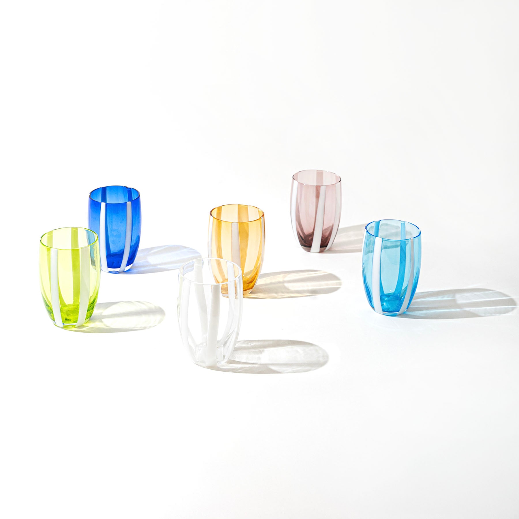 Cinque bicchieri di vetro colorato con strisce bianche verticali, disposti su sfondo bianco con ombre nette, colori giallo, blu, arancione, rosa e