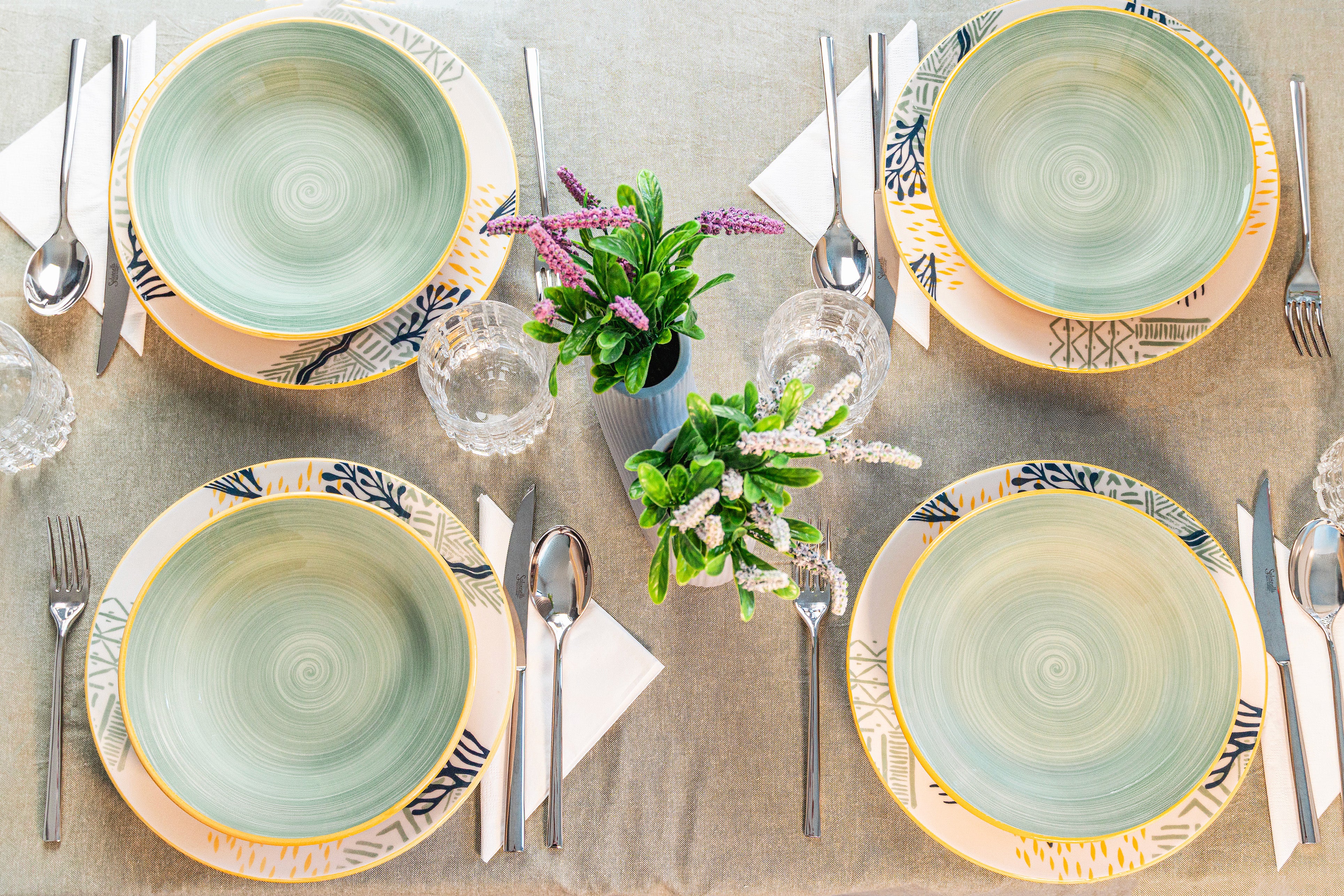 Quattro piatti decorati in ceramica, posati su una tovaglia chiara, con posate e bicchieri trasparenti. Due piccoli vasi con piante verdi e fiori.