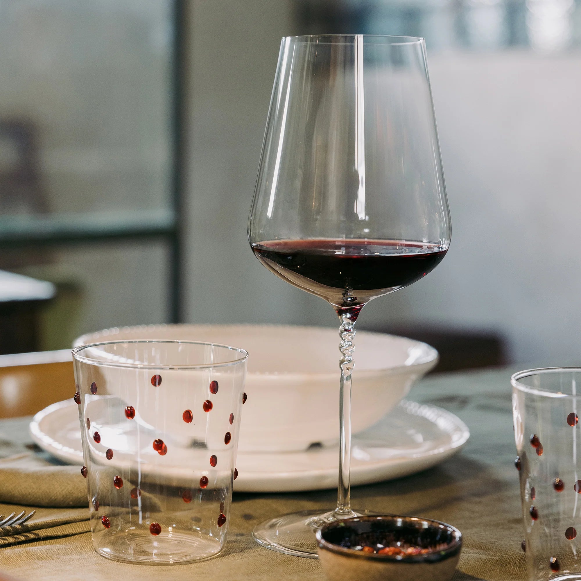 Calice di vetro trasparente con gambo intrecciato contenente vino rosso, bicchieri trasparenti con puntini rossi, piatti bianchi e ciotola su tavolo