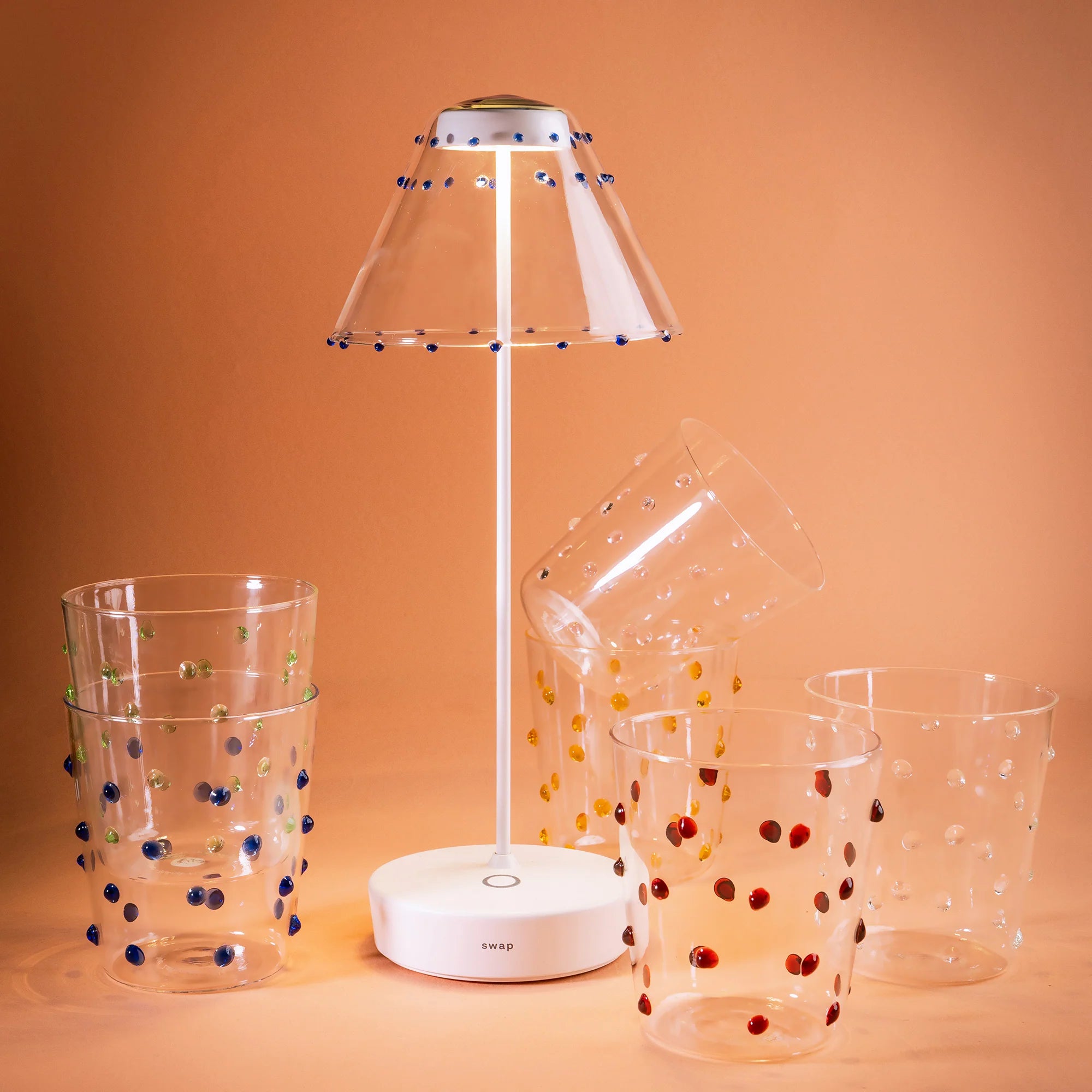 Lampada da tavolo bianca con paralume trasparente e decori blu, circondata da cinque bicchieri trasparenti con puntini colorati, sfondo arancione.