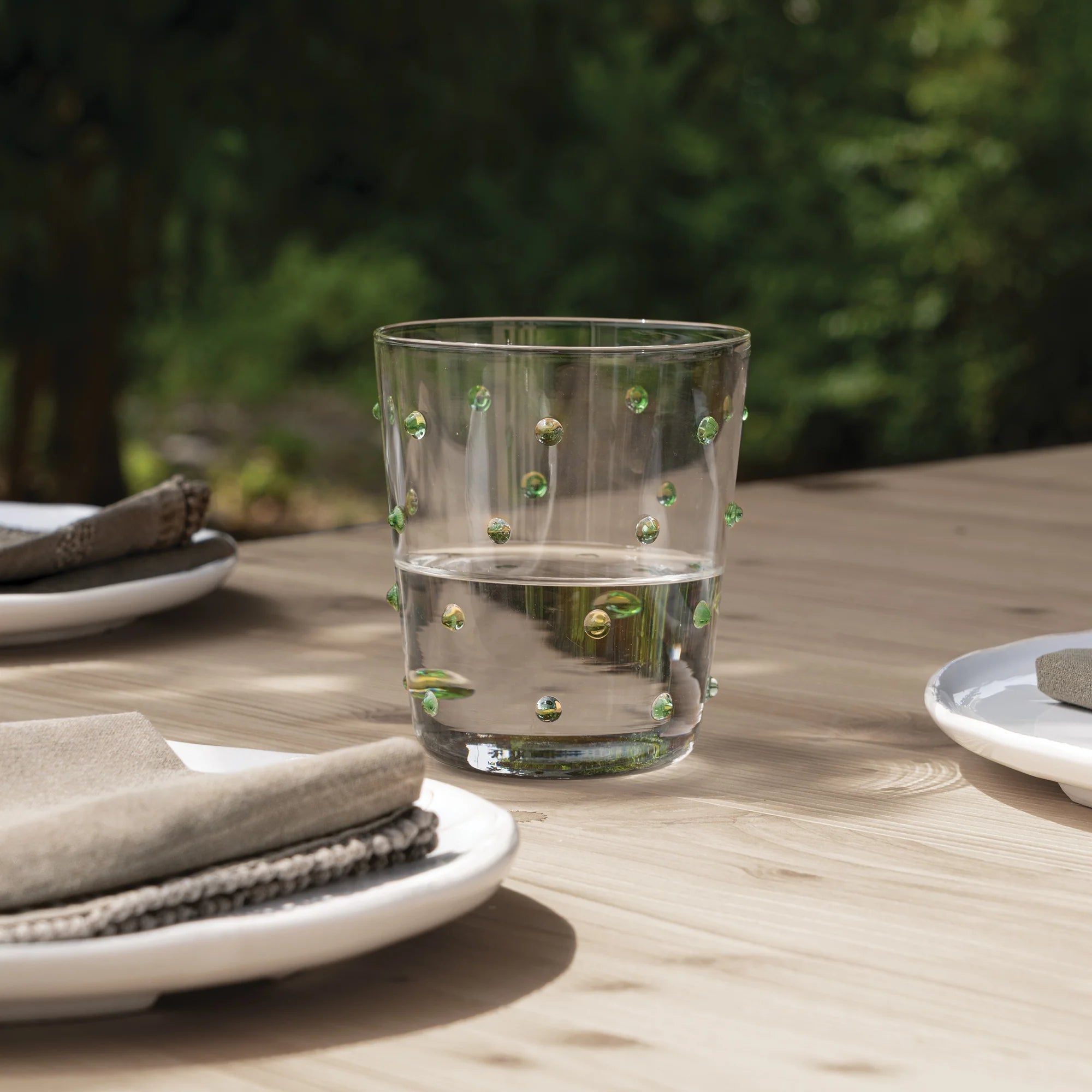 Bicchiere trasparente con piccoli decori verdi a rilievo, parzialmente pieno d'acqua, su tavolo di legno chiaro con piatti bianchi e tovaglioli