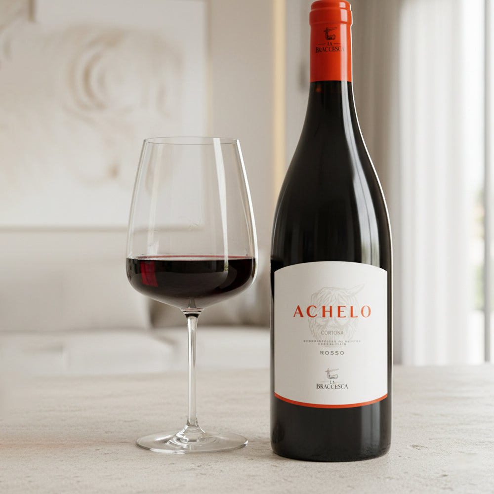 Bottiglia di vino rosso Achelo con etichetta bianca e testo arancione accanto a calice di vetro trasparente con vino rosso versato, su superficie