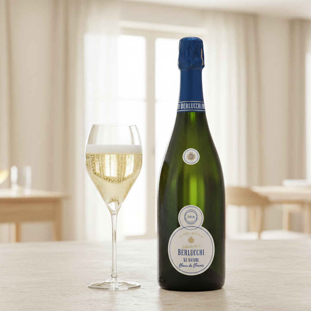 Bottiglia di champagne Berlucchi 2016 accanto a un calice con spumante dorato, su un tavolo con sfondo luminoso e arredo semplice.