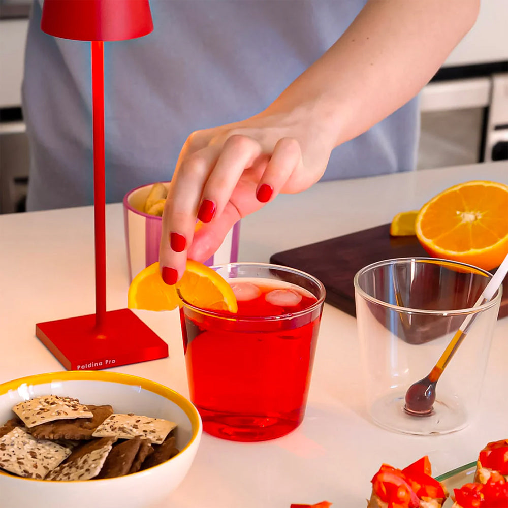 mano con unghie rosse che poggia una fetta d'arancia su bicchiere con bevanda rossa e ghiaccio su tavolo bianco in cucina
