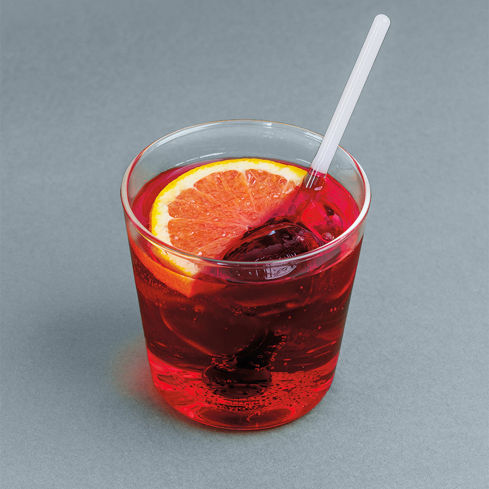 Bicchiere trasparente con aperitivo rosso, fetta di arancia, due cubetti di ghiaccio e cannuccia bianca su sfondo grigio.