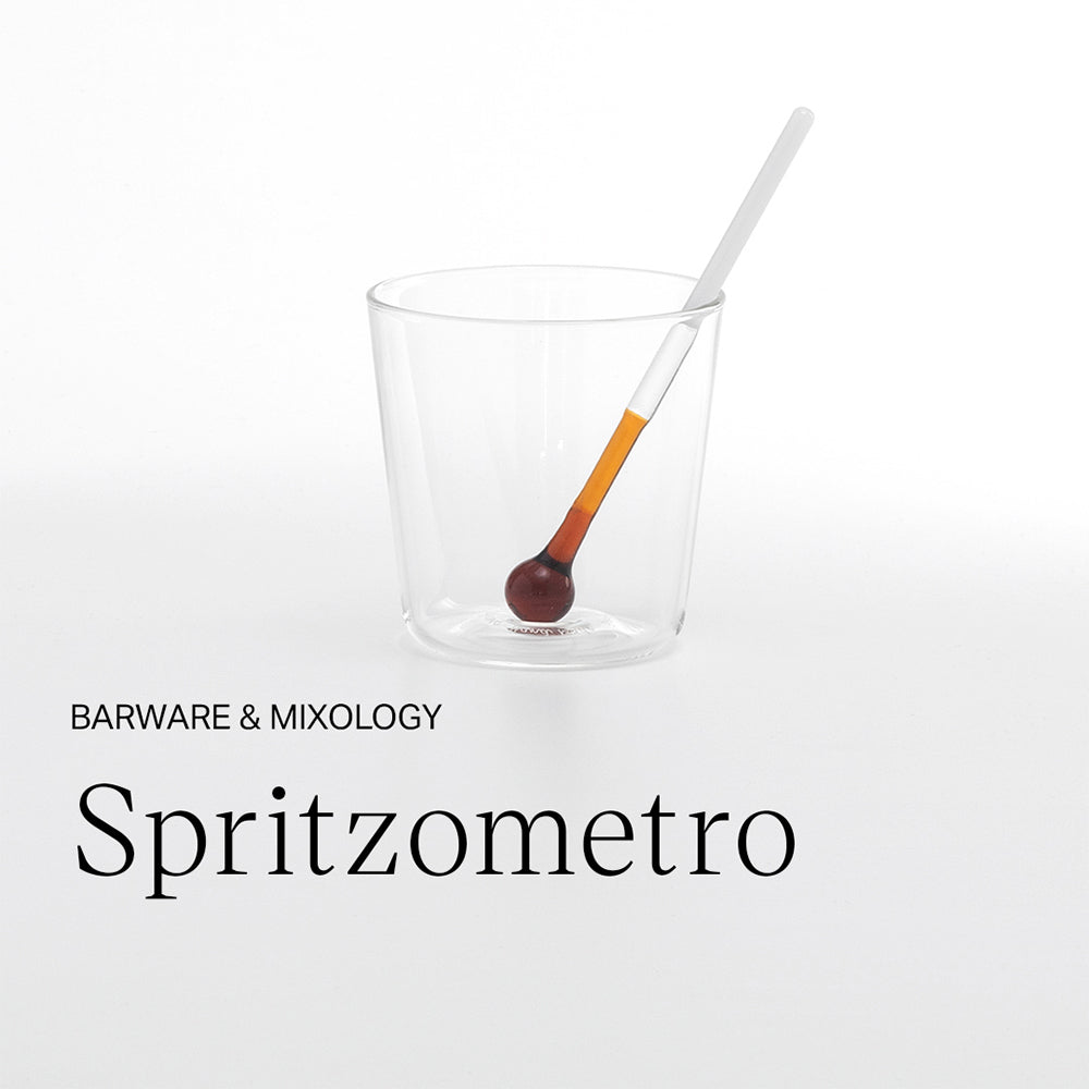 Bicchiere trasparente in vetro con bacchetta dosatrice con estremità tonda marrone e arancione su fondo bianco.