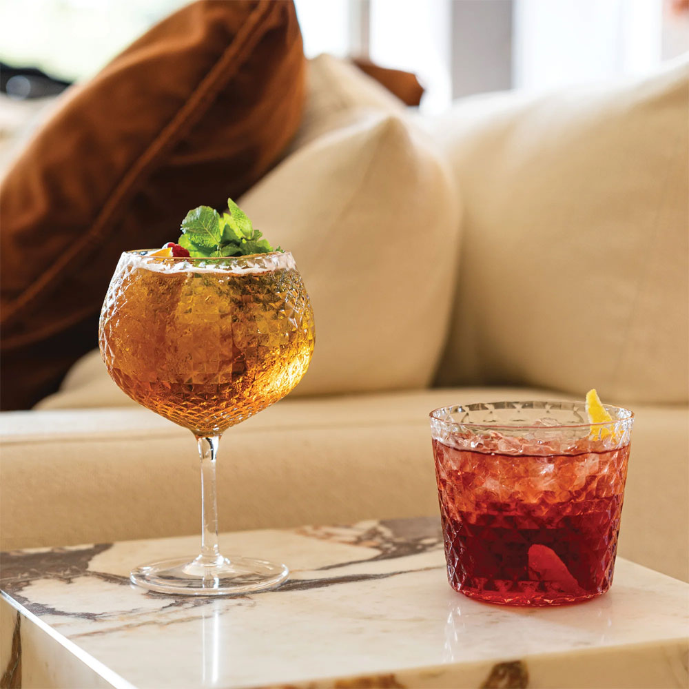 Due bicchieri trasparenti con superficie sfaccettata, contenenti drink giallo ambrato e rosso con ghiaccio e garnish, su tavolo marmo.