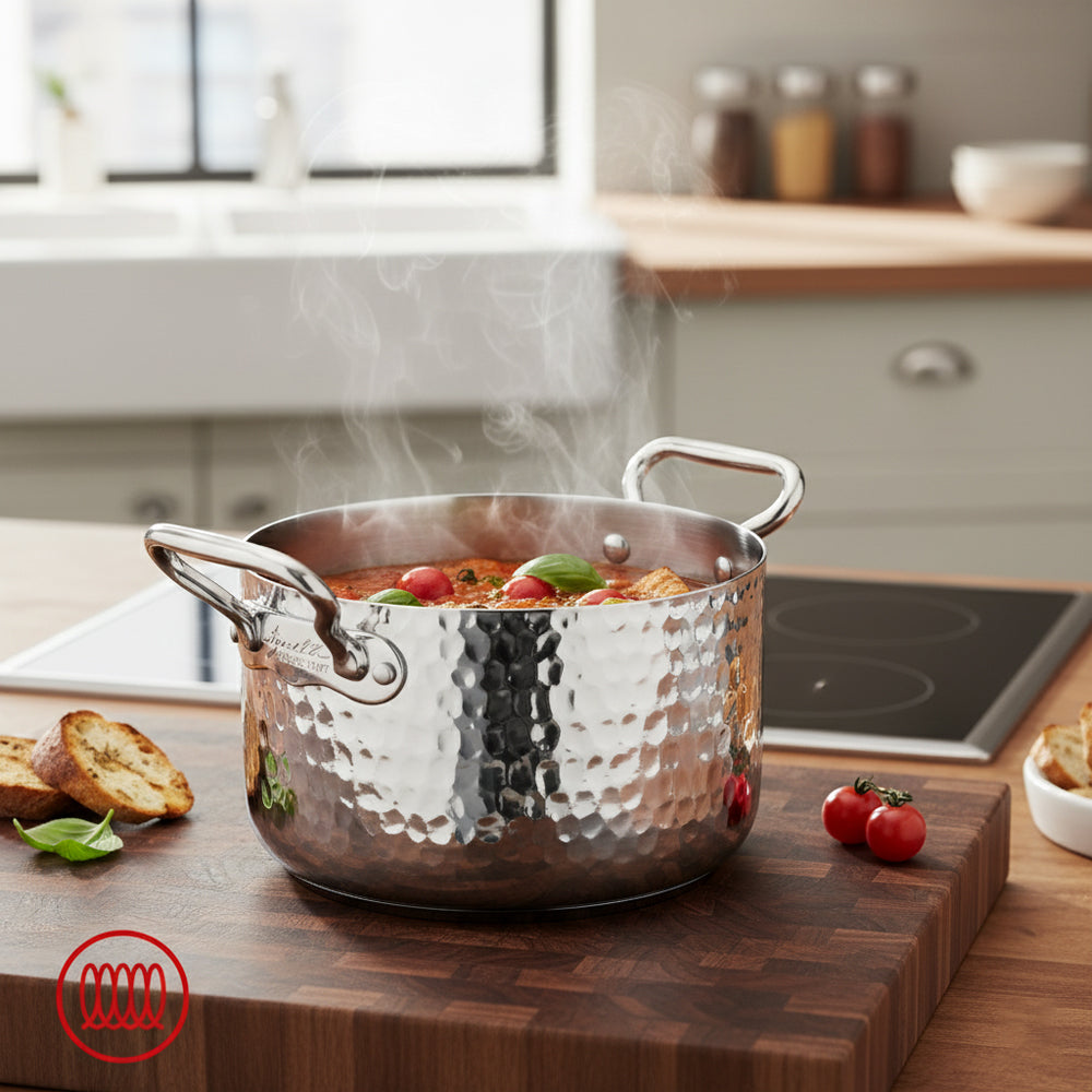 Pentola in acciaio inox martellato con due manici pieni di sugo caldo con pomodorini e basilico su tagliere di legno in cucina moderna.