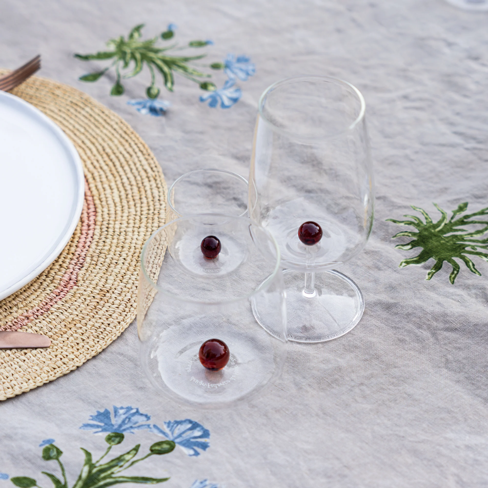 Tre bicchieri di vetro trasparente con base rossa sferica, su tovaglia beige con decorazioni floreali azzurre e verdi e tovaglietta in