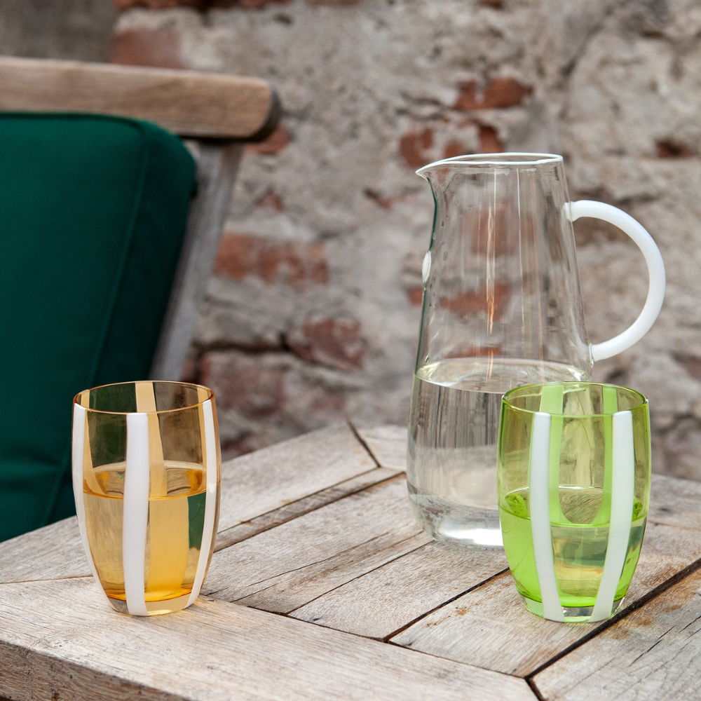 Due bicchieri in vetro colorato con decorazioni bianche su tavolo legno chiaro, brocca d'acqua trasparente e sfondo muro pietra e sedia