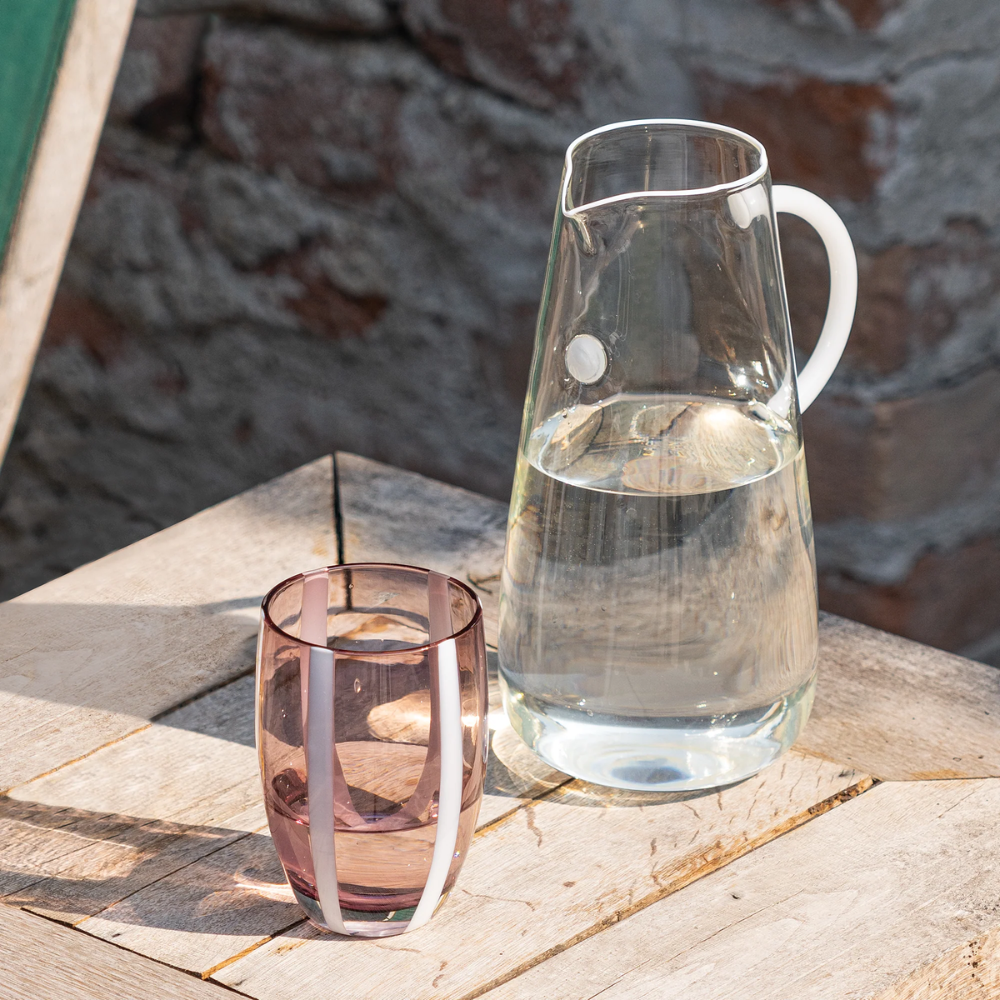 caraffa in vetro trasparente con acqua e bicchiere viola con strisce bianche su tavolo di legno chiaro all'aperto contro muro scuro di