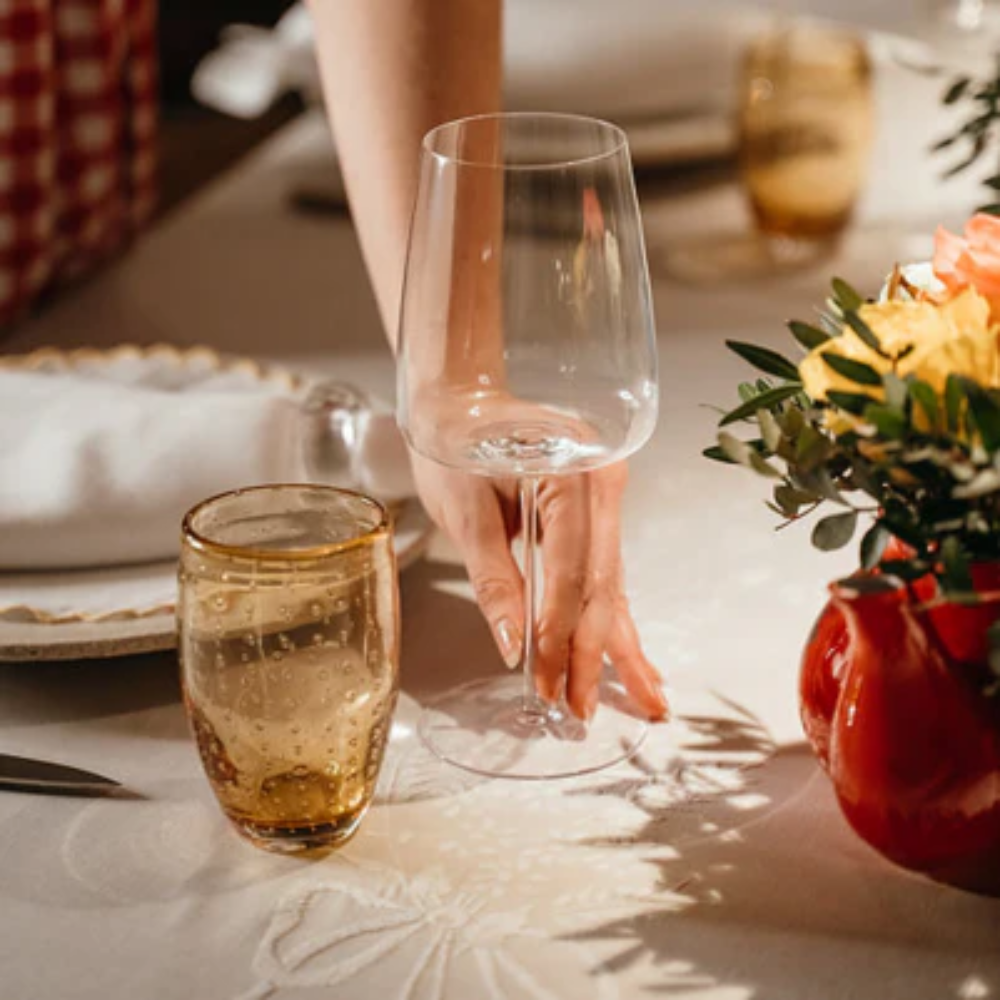 Calice da vino trasparente affiancato a bicchiere di vetro ambra con bolle, mano che prende il calice, bouquet fiori gialli e rossi