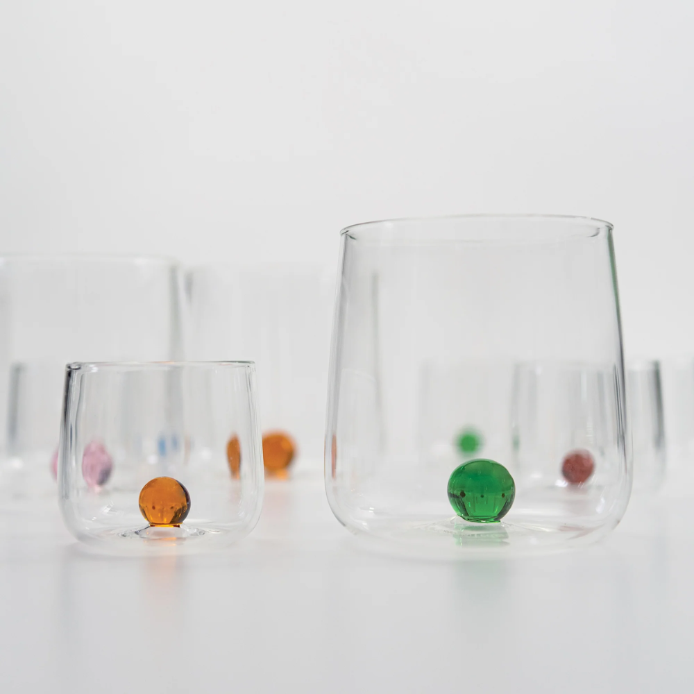 Set di bicchieri di vetro trasparente con sfere colorate inserite sul fondo, su superficie bianca e sfondo chiaro.