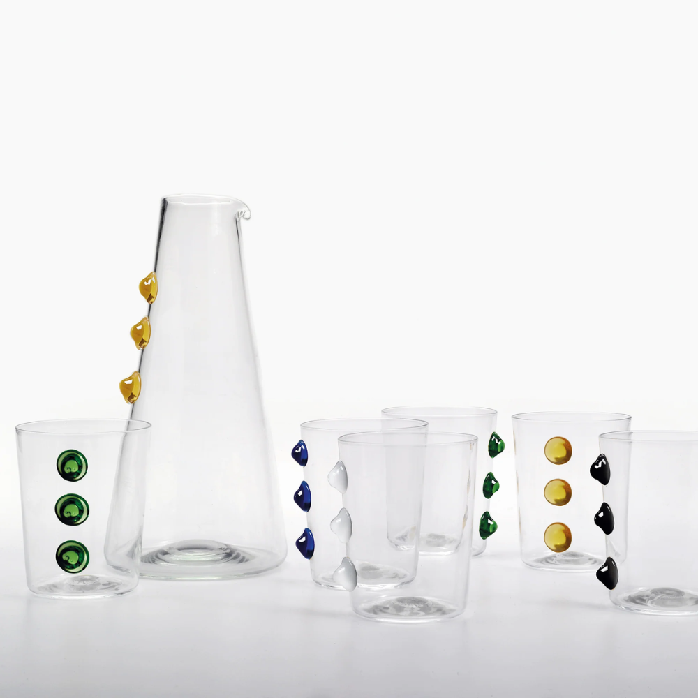 Caraffa trasparente e sei bicchieri in vetro con borchie colorate verde, giallo, blu, bianco e nero su sfondo bianco.