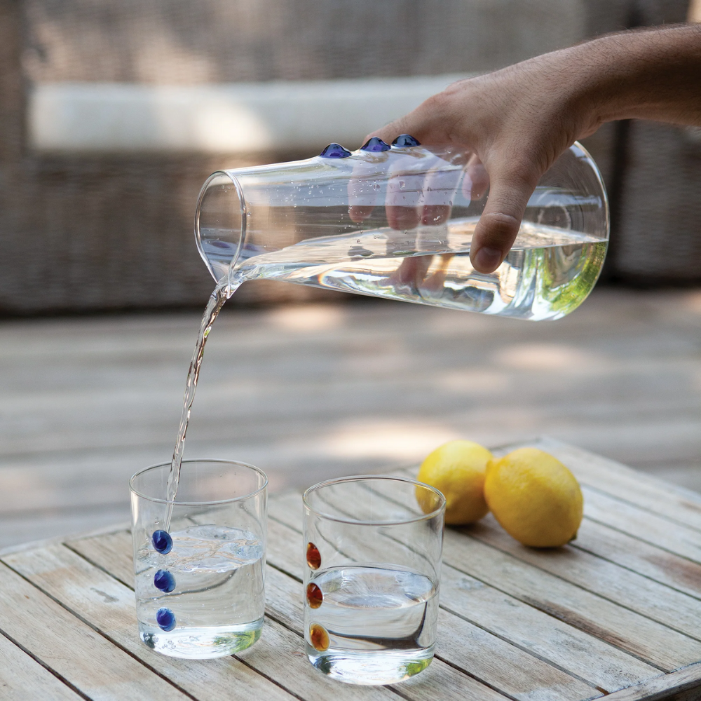 Brocca di vetro trasparente con decorazioni blu versata in due bicchieri con elementi colorati su tavolo in legno all'aperto con limoni.
