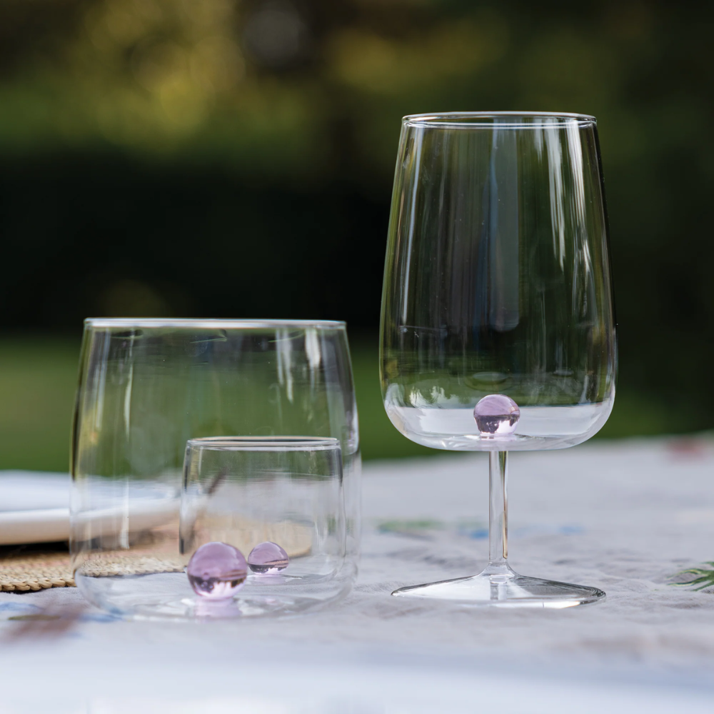 due bicchieri trasparenti in vetro, uno tumbler con tre sfere rosa sul fondo e uno calice con sfera rosa, su tovaglia bianca all'aperto