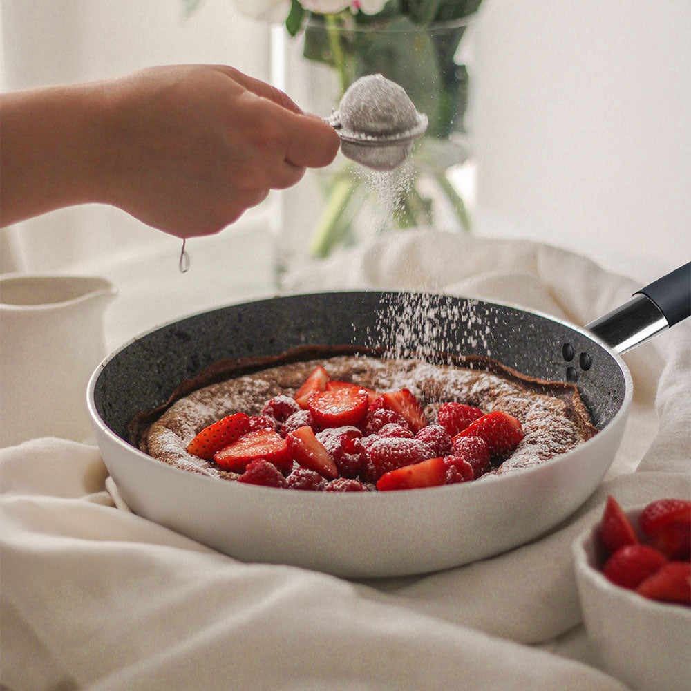 Padella grigia con manico nero contenente dolce con fragole e lamponi, zucchero a velo spolverato da mano con colino metallico, sfondo