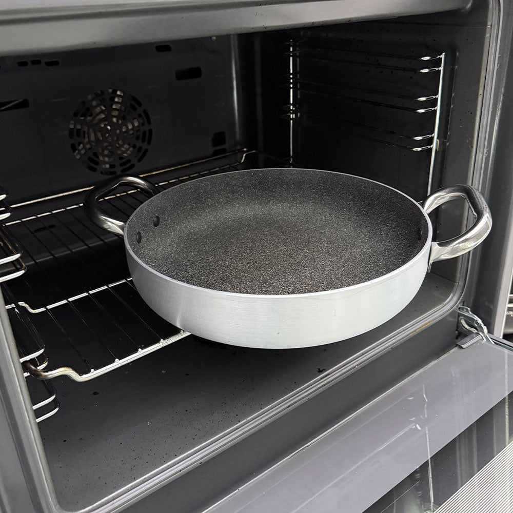 Padella rotonda con doppio manico in metallo lucido e interno antiaderente nero con effetto pietra, dentro forno aperto grigio.