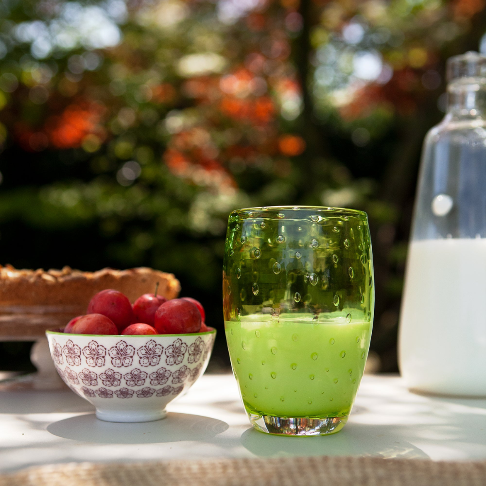 Bicchiere verde in vetro con bolle contenente liquido verde, ciotola bianca con prugne rosse e bottiglia di latte in ambiente esterno