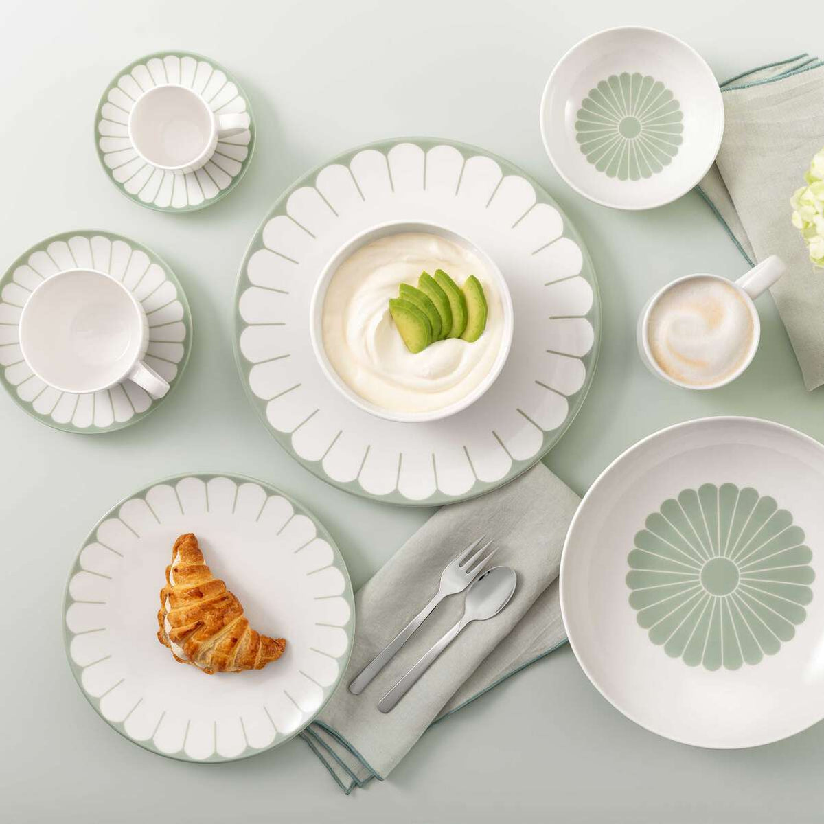 Set di stoviglie bianche e verdi con motivi ad arco, contenente caffè, croissant e yogurt con avocado, posate e tovagliolo su tavolo chiaro.