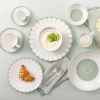 Set di stoviglie bianche e verdi con motivi ad arco, contenente caffè, croissant e yogurt con avocado, posate e tovagliolo su tavolo chiaro.