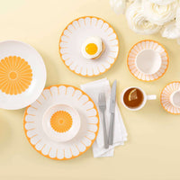 Set da tavola con piatti bianchi e arancioni, posate grigie, una tazza di tè e un piatto con un uovo all'occhio di bue su un sottofondo giallo.