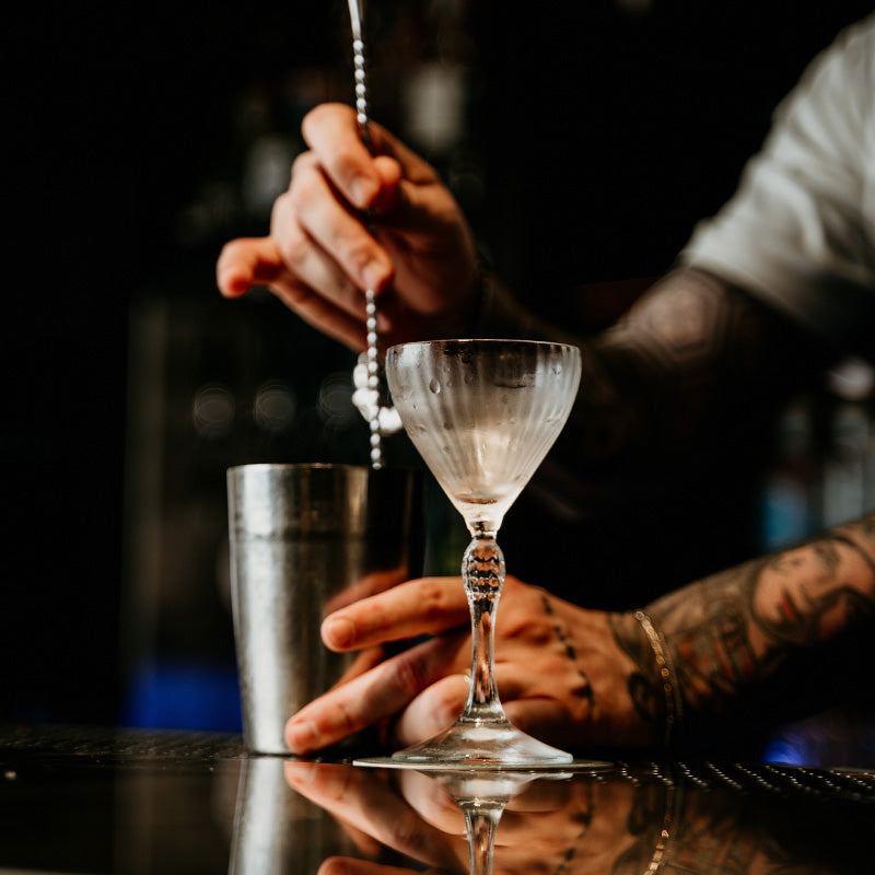 Calice di vetro con gocce di condensa su un bancone riflettente, lampada da cocktail in metallo e mano tatuata che mescola un drink.
