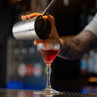 mano con tatuaggi versa liquido rosso da shaker metallico su calice di vetro trasparente in bar sfocato