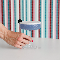 mano con smalto rosa che tiene calice di vetro con cocktail viola con schiuma bianca e mora su sfondo a righe colorate
