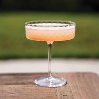 Calice trasparente con superficie sfaccettata, contenente un cocktail rosa tenue, appoggiato su tavolo in legno, sfondo sfocato verde.