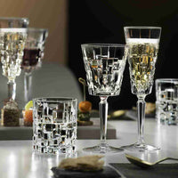 Set di bicchieri in vetro trasparente con motivi geometrici, composto da calici e tumbler su tavolo con sfondo scuro e antipasti sfocati.