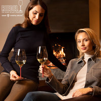 due donne con abiti autunnali tengono bicchieri di vino bianco con gambo rosso davanti a un camino acceso in ambiente accogliente e moderno
