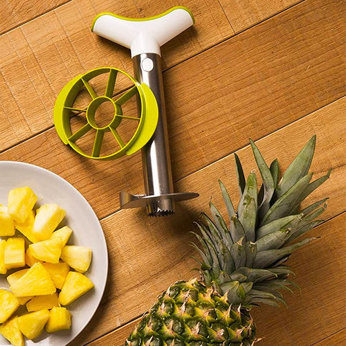 Strumento per tagliare ananas in acciaio inox con manico bianco e verde, accanto a fette di ananas su piatto e ananas intero.
