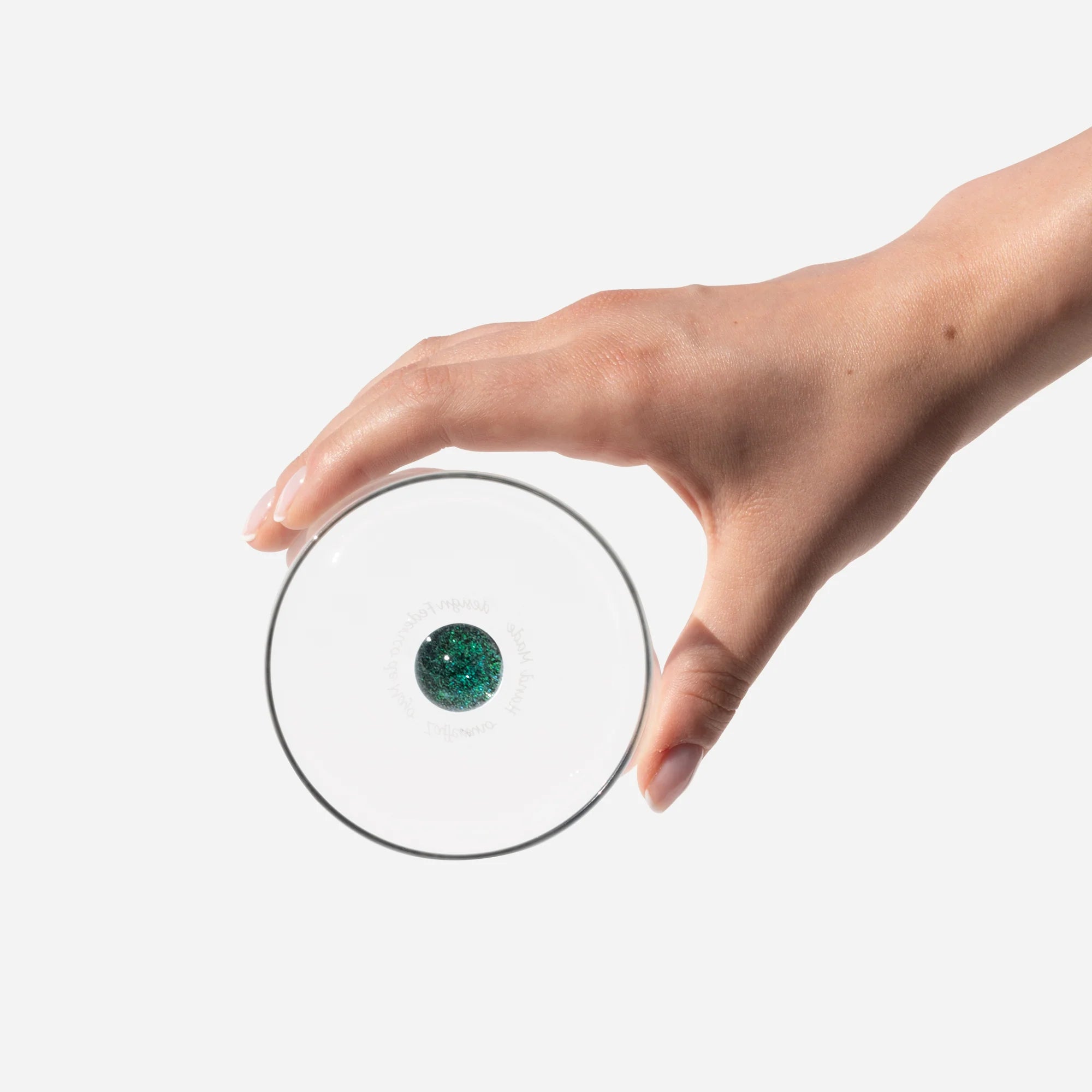mano femminile con unghie curate che regge un bicchiere trasparente con palla verde brillante al centro, sfondo bianco