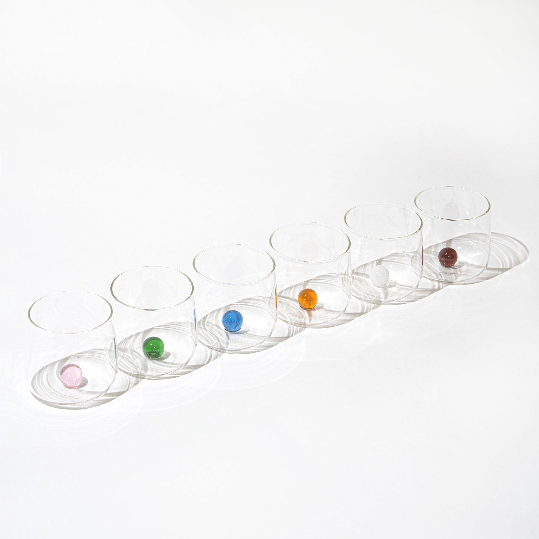 Set di sette bicchieri trasparenti in vetro, ognuno con una biglia colorata diversa all'interno, su sfondo bianco.