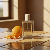 Bottiglia trasparente di profumo colore giallo chiaro con tappo crema accanto a un'arancia con scorza arrotolata su piastrelle beige e marroni, luce