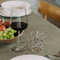 Bicchiere da vino trasparente con vino rosso e bicchiere trasparente decorato da puntini rossi su tovaglia verde con ciotola di frutta e piatti