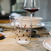 Bicchiere trasparente in vetro con pois rossi appoggiato su tavola imbandita con piatti beige e calice di vino rosso sullo sfondo.